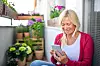 Eldre kvinne i rød cardigan bruker smarttelefon på en balkong omgitt av grønne planter.