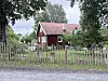 Rødt trehus med hvite detaljer bak et værslitt tregjerde og en grusvei.