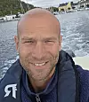 Smilende mann i redningsvest på båt med sjø og hus i bakgrunnen.