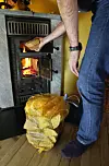 Person legger ved i en vedovn med synlige flammer og en sekk med ved på gulvet.
