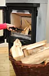 Hånd som tenner ved i en svart vedovn, med en flettet kurv full av ved i forgrunnen