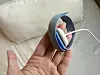 Hånd holder sammenfoldede blå hodetelefoner med hvit kabel foran en lys pute.