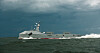 Grått marinefartøy seiler gjennom bølger under mørk, skyet himmel.