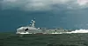 Grått marinefartøy seiler gjennom bølger under mørk, skyet himmel.