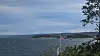 Norsk flagg ved vannet, med skogkledd kystlinje og overskyet himmel i bakgrunnen.