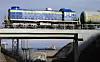 Blått og gult lokomotiv med tankvogn på jernbanebro over industriområde ved havn.