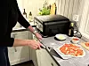 Person fordeler topping på pizzabunn på bakepapir ved siden av en svart benkeovn.