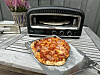 Nystekt pizza på bakepapir foran en liten elektrisk pizzaovn på et grått utebord.