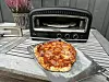 Nystekt pizza på bakepapir foran en liten elektrisk pizzaovn på et grått utebord.