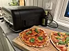 To nystekte pizzaer på skjærebrett ved siden av en svart pizzaovn på kjøkkenbenken.
