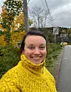 Kvinne i gul strikkegenser ved vei med 30-skilt, høstløv og boliger i bakgrunnen.