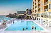 Gjester i utendørsbasseng ved et kysthotell, med hav, solsenger og hotellbygning.