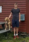 Mann står ved en hund på en benk utenfor en rød trebygning.
