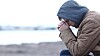 Person med lue og tykk jakke sitter sammenkrøket ved et kaldt vann.
