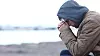 Person med lue og tykk jakke sitter sammenkrøket ved et kaldt vann.
