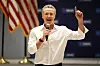 Gavin Newsom taler med mikrofon og hevet pekefinger foran amerikansk flagg og blå bakgrunn.