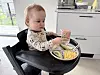 Småbarn i barnestol spiser mais, avokado og brød fra en delt tallerken på kjøkkenet.