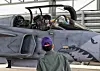 Pilot i cockpit på grått jagerfly med haimaling, med en person foran flyet.