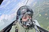 Pilot med hjelm og oksygenmaske i cockpiten på et jagerfly over landskap.