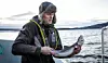 Mann i vinterklær holder en fisk på dekk på en båt, med fjord og fjell i bakgrunnen.