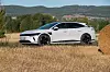 Hvit Kia EV6 parkert på tørr åker ved en rundball, med skogkledde åser i bakgrunnen.