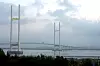 Lang hengebro over vann under skyet himmel, med busker i forgrunnen.