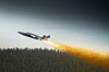 Jetfly flyr over skog med lang ild- og røykstripe bak seg.