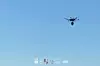 Drone svever mot klar blå himmel, med små logoer og tekst nederst i bildet.