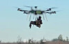 Stor drone flyr med en person hengende i seler under, over trær og blå himmel.