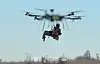 Stor drone flyr med en person hengende i seler under, over trær og blå himmel.