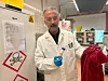 Mann i laboratoriefrakk og blå hansker holder et prøveglass ved fareskilt.