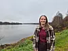 Kvinne i rutete jakke står på en gresskledd elvebredd ved stille vann.