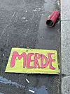 Gult skilt med teksten «MERDE» på fortau ved rød kopp og sigarettstumper.