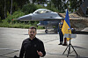 Volodymyr Zelenskyj taler foran et F-16-kampfly, ukrainsk flagg og en uniformert vakt.