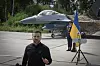 Volodymyr Zelenskyj taler foran et F-16-kampfly, ukrainsk flagg og en uniformert vakt.