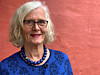 Eldre kvinne med briller og blå jakke foran en rosa vegg.