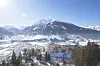 Hvitt slott i en snødekt alpelandsby, omgitt av skog og fjell under klar blå himmel.