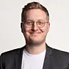 Mann med runde briller, mørk blazer og hvit T-skjorte mot lys bakgrunn.
