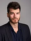 Mann med skjegg og mørk blazer i studioportrett mot grå bakgrunn.