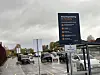 Biler på parkeringsplass med informasjonsskilt og høyblokk i bakgrunnen.