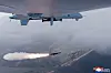 Militær drone over skyer mens et missil med flammehale avfyres over et kystlandskap.