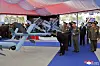 Uniformerte tjenestemenn ved grå droner og militært utstyr under en overbygget utstilling.