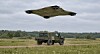 Militært terrengkjøretøy på et felt under en stor drone i lav høyde.