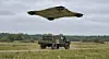 Militært terrengkjøretøy på et felt under en stor drone i lav høyde.