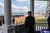 Mann i mørk dress ser ut over en oppstilt folkemengde og et seremoniområde fra en søylegang.