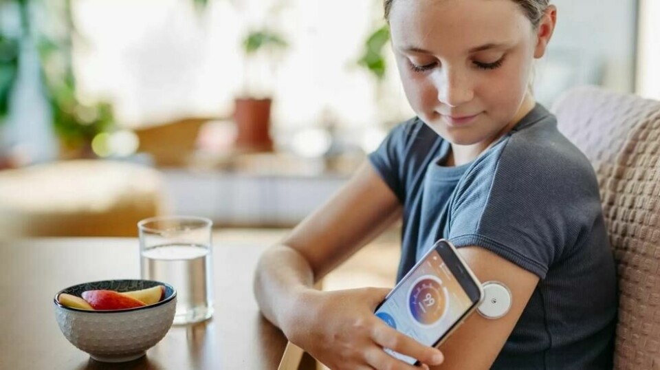 Jente som følger med på sitt eget blodsukker på en mobilapp. Hun sitter ved bordet. Hun har et glass vann og en skål med frukt foran seg. Totalt har rundt 3.500 norske barn og unge diabetes i Norge. Ny studie viser at de har fått et mer stabilt blodsukker, mye takket være ny teknologi.
