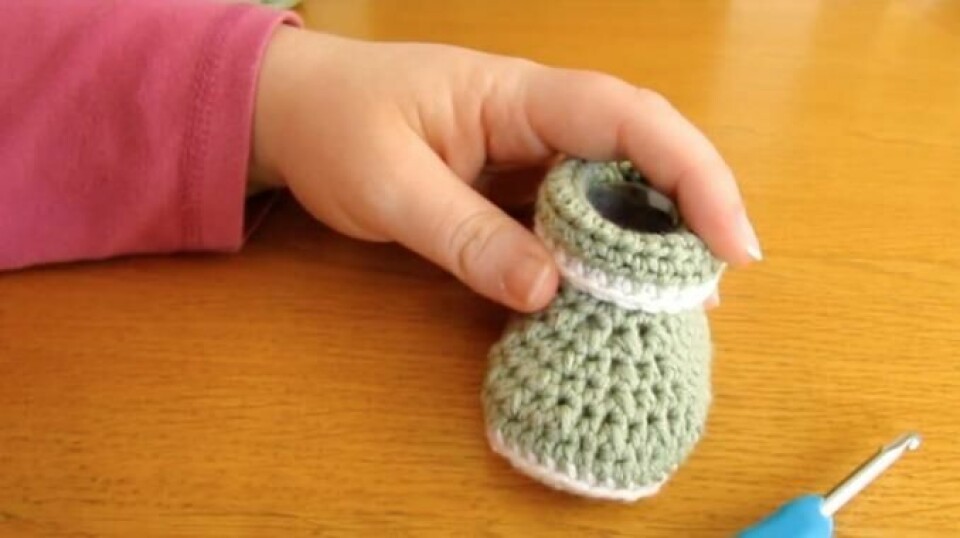 Super video av hekleoppskrift for nybegynnere, på babytøfler. Disse hekler du på 1-2-3! (Foto: Wooly Wonders Crochet) Bilde av person som er ferdig å hekle, med ferdige tøfler fra en super hekleoppskrift for nybegynnere. Grønne og hvite babytøfler i hand på trebord.