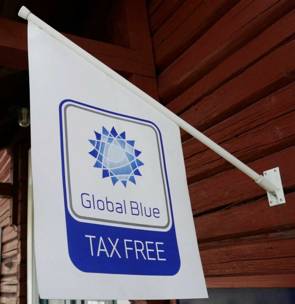 Se etter butikker med et slikt skilt eller flagg for å vite hvor du kan handle tax free. Bilde av et hvitt flagg som henger på en rød vegg. Flagget har blå skrift hvor det står tax free. Vi forklarer hvordan du kan få momsen tilbake når du handler i utlandet.