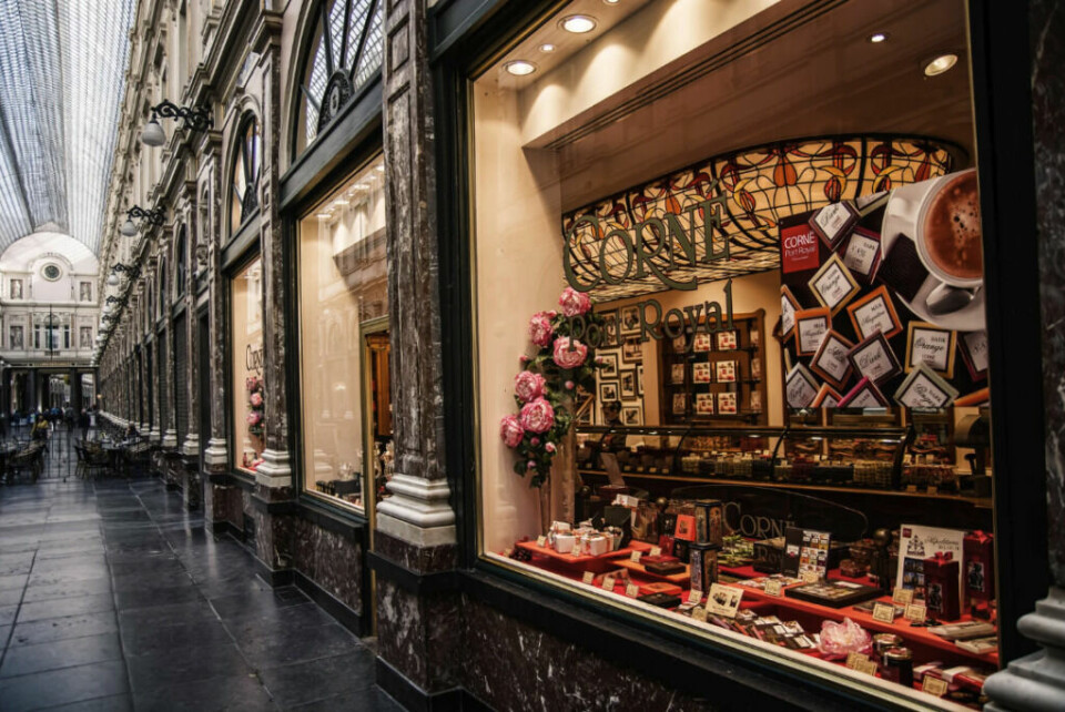 Belgia er kjent for både vaflene, ølen og sjokoladen sin. Corne Port-Royal, er en eksklusiv sjokoladeforretning som tilbyr utsøkte belgiske sjokolader, og i bakgrunnen ser vi Galeries Royales Saint-Hubert, et ikonisk shoppingområde i Brussel med en elegant glasstakpassasje. (Foto: Canva) Dette bildet viser en elegant butikkfasade i et luksuriøst kjøpesenter, sannsynligvis Galeries Royales Saint-Hubert i Brussel, Belgia. Butikken, Corne Port-Royal, er en eksklusiv sjokoladeforretning som tilbyr utsøkte belgiske sjokolader, vakkert presentert i et klassisk interiør. Detaljer i bildet: Fasaden har store vinduer med forseggjorte, marmorlignende søyler og belysning som gir en varm, innbydende glød. Et dekorativt glassmaleri over butikken gir et vintage og sofistikert preg. Butikkutstillingen viser luksuriøse sjokoladeesker, roser og delikate gaver, noe som understreker butikkens eksklusive preg. I bakgrunnen ser vi den flotte arkitekturen i Galeries Royales Saint-Hubert, et ikonisk shoppingområde i Brussel med en elegant glasstakpassasje, hvor folk kan spasere og nyte eksklusive butikker og kafeer. Bildet fanger en klassisk europeisk shoppingopplevelse, med en blanding av luksus, historie og kultur som Brussel er kjent for.