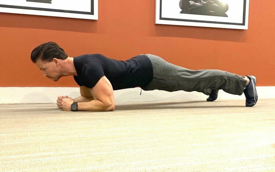 Planken: Begynn liggende på magen med hendene under skuldrene. Legg så hele vekten av kroppen på underarmene ved å stramme hele kroppen og løft deg opp i en rett linje. Hold her så lenge du makter. (Foto: Cornelis Elander) Bilde av personlig trener Cornelis Elander som trener diagonalstrekk på treningsmatte. Han har på seg en svart t-skjorte og en grå treningsbukse, og i bakgrunnen på bildet ser vi en oransje vegg med bilder. Han befinner seg i et treningsrom. Dette er en styrkeøvelse med fordeler for mye av kroppen og deriblandt bein, core, magemuskler, armer og rumpe, og en av styrkeøvelsene i vårt 30-dagers gratis treningsprogram og styrkeprogram.